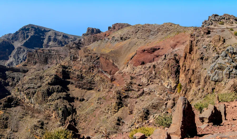 El precipicio de la Caldera de Taburiente al oeste del Roque de los Muchachos: la crestería occidental con sus estratos volcánicos rojizos y ocres y la senda del cordal asomando por el borde, La Palma